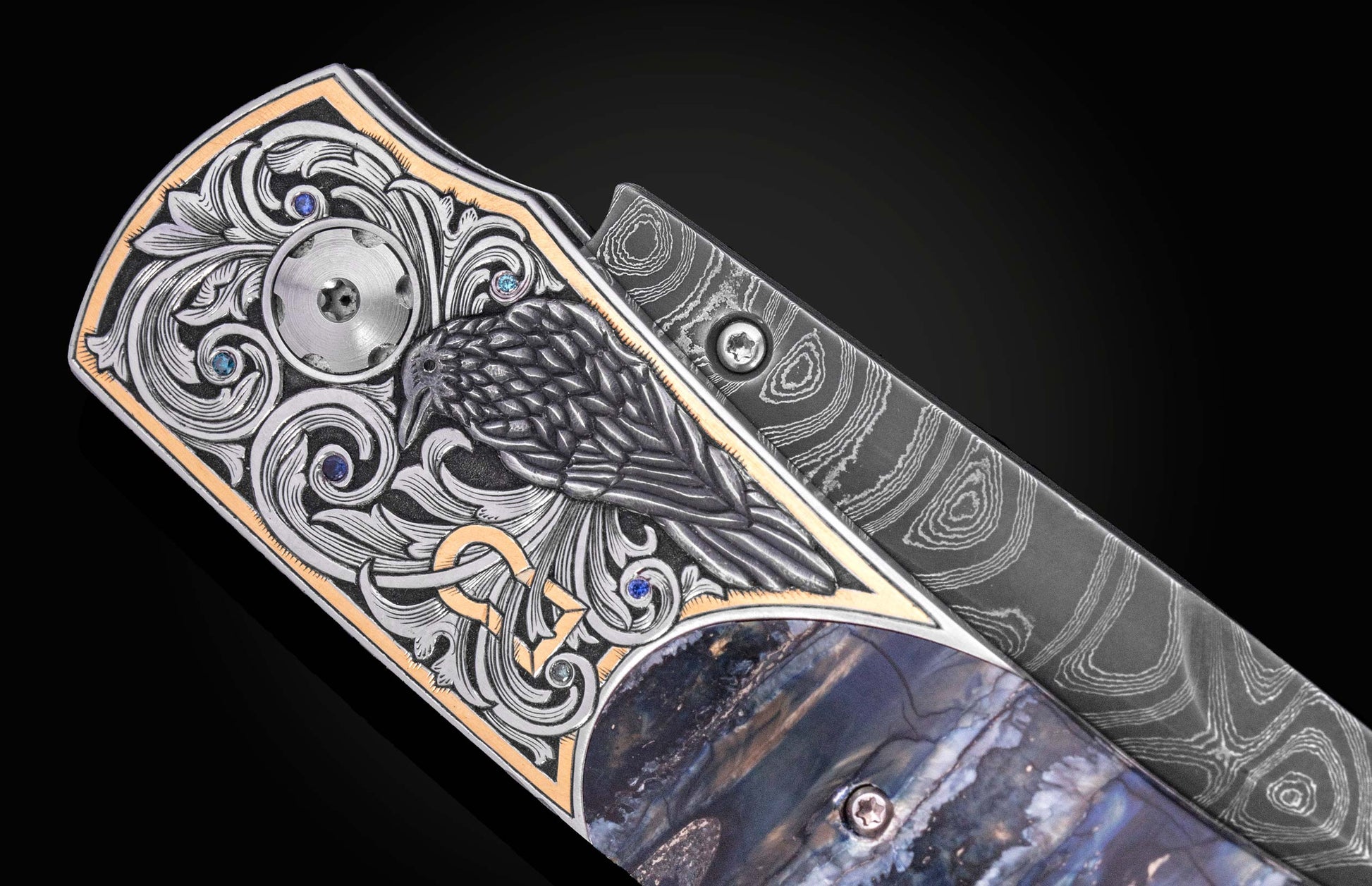 Luxury folding knife with intricate raven engraving and sapphire accents, featuring damascus steel blade and marbled handle, side view close-up