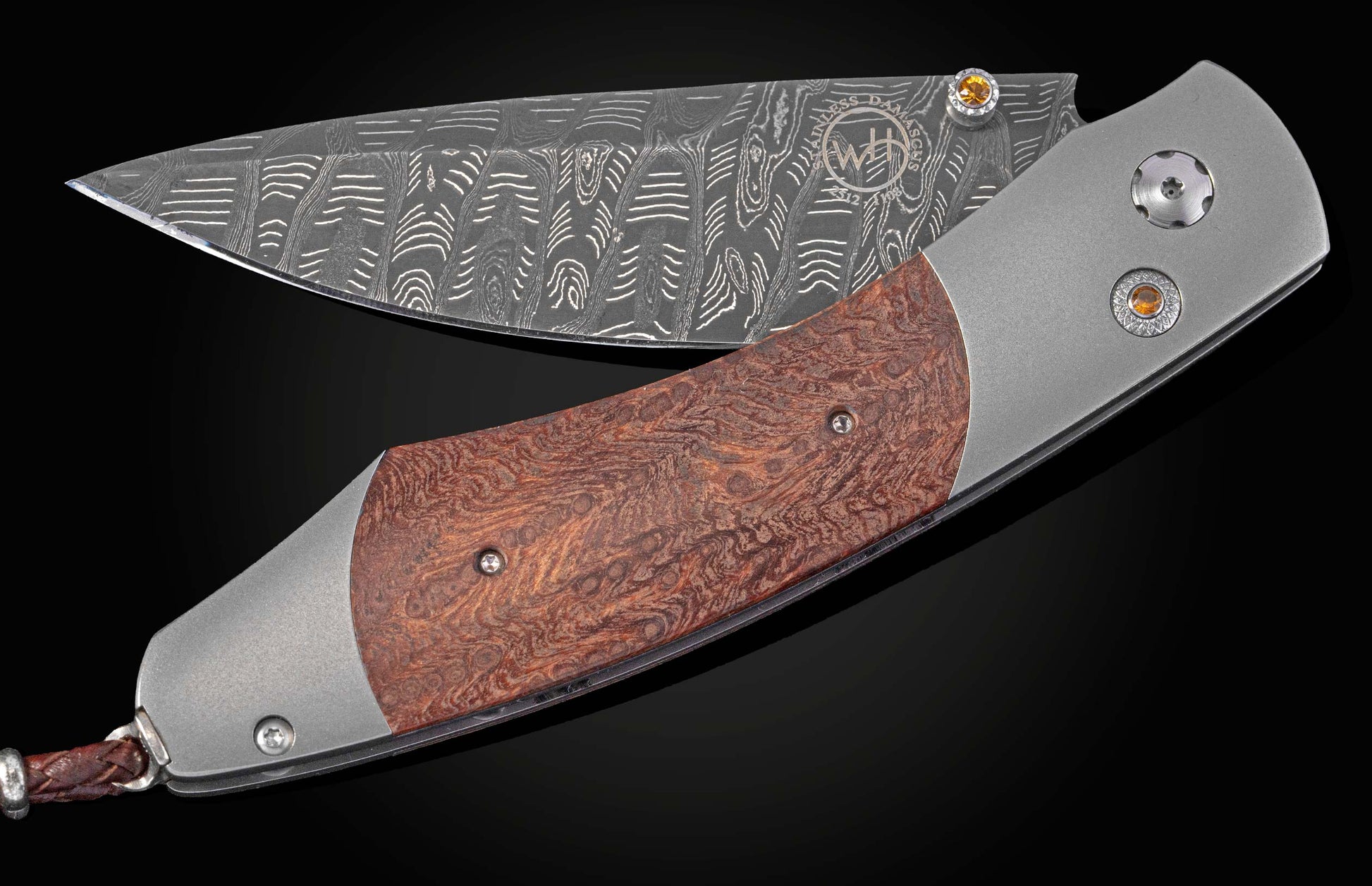 Side view of a folding knife featuring a patterned Damascus blade, reddish burl wood handle, amber gems, and a braided leather lanyard.