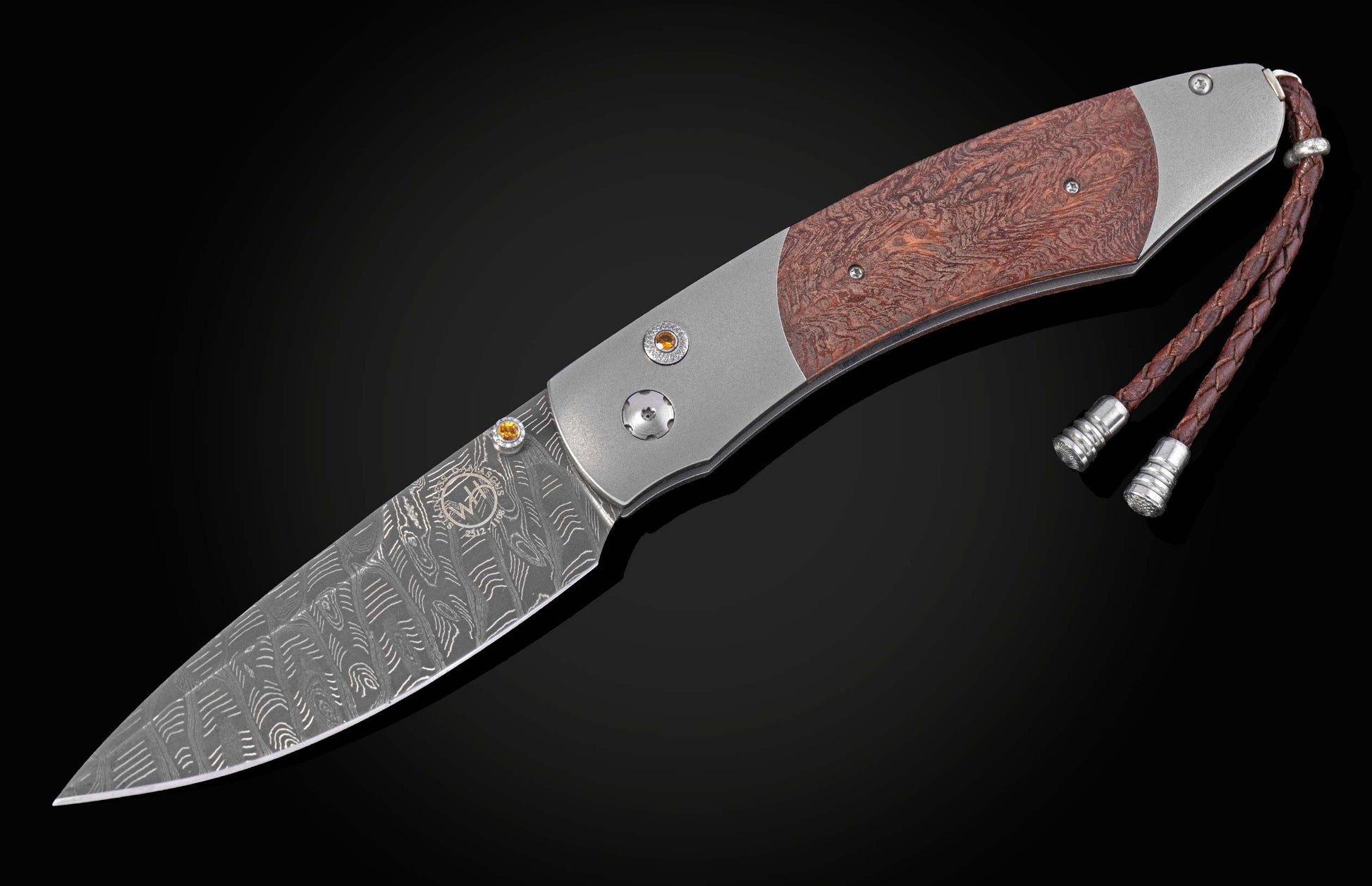 Folding knife with a Damascus steel blade, redburl wood handle, orange gemstone accents, and a braided leather lanyard.
