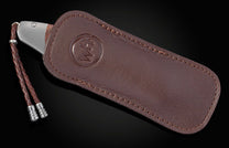Knife with a metal end and dark handle, partially inserted into a brown leather sheath with an embossed logo, featuring a braided lanyard with metal tips.