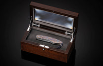 Decorative folding knife with a colorful, intricately patterned handle and a Damascus blade, resting in an open dark wood display box.