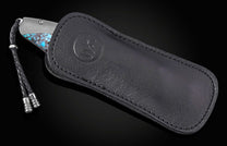 Luxury folding knife with turquoise inlay handle by B12, partially sheathed in black leather case with embossed logo and braided cord.