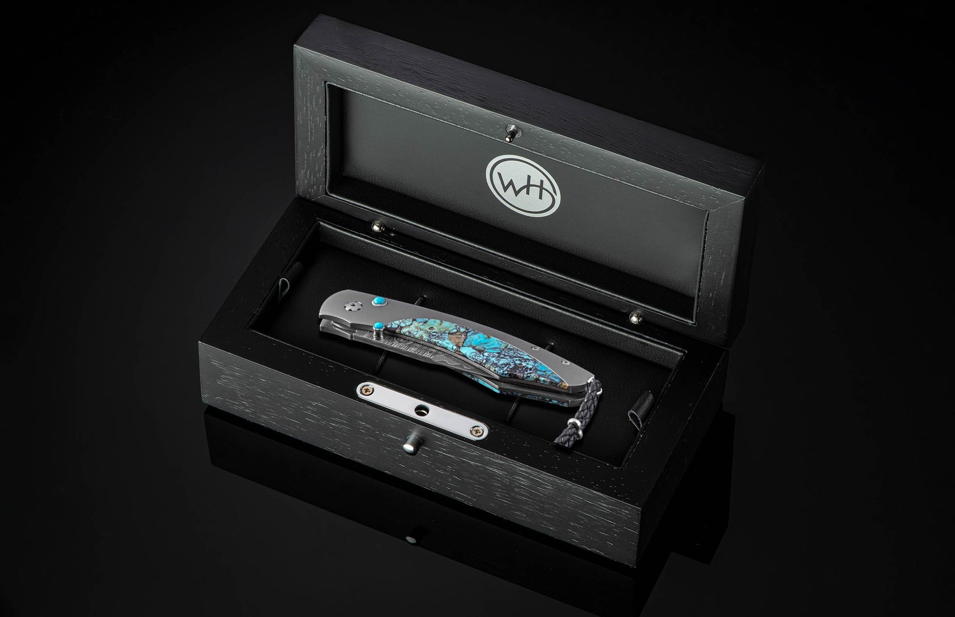 Turquoise-stone inlaid folding knife by B12, displayed in black textured presentation box with silver hardware and braided lanyard, top-down view.