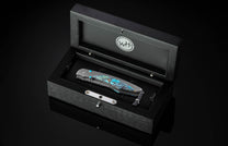 Turquoise-stone inlaid folding knife by B12, displayed in black textured presentation box with silver hardware and braided lanyard, top-down view.