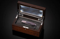 An intricate folding knife with a patterned handle and dark blade, presented in an open dark wood display box with a glass lid.