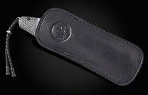 Silver patterned blade knife in black leather sheath with embossed logo and braided lanyard, side view