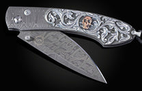 Partially open Damascus steel folding knife with ornate silver handle engraving featuring a rose gold skull.