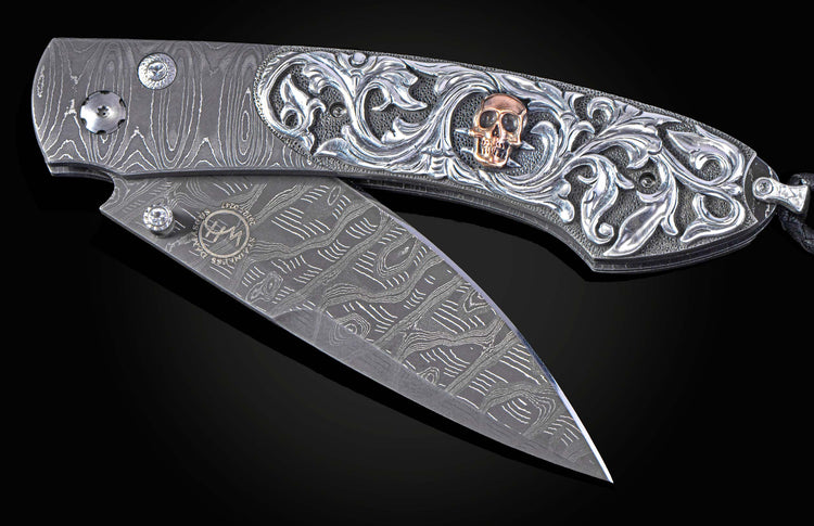 Partially open Damascus steel folding knife with ornate silver handle engraving featuring a rose gold skull.