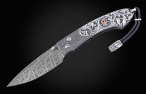 Damascus steel folding knife with ornate silver handle engraving featuring a copper skull and a black braided leather lanyard.