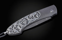Ornate knife with a silver skull and floral engraving, Damascus steel handle, and black braided leather cord.