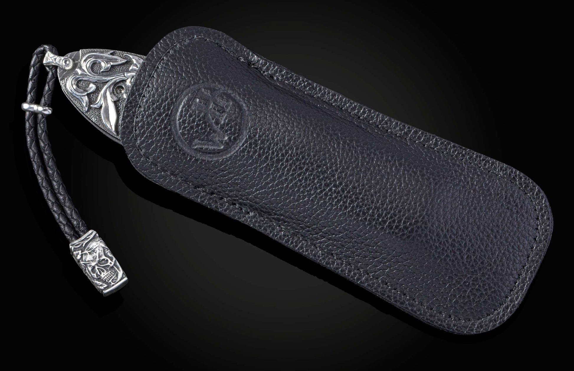 Decorative knife with an ornate silver-engraved handle, partially tucked into a black leather sheath with an embossed logo and a skull lanyard.