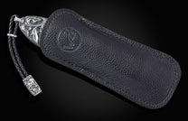 Decorative knife with an ornate silver-engraved handle, partially tucked into a black leather sheath with an embossed logo and a skull lanyard.