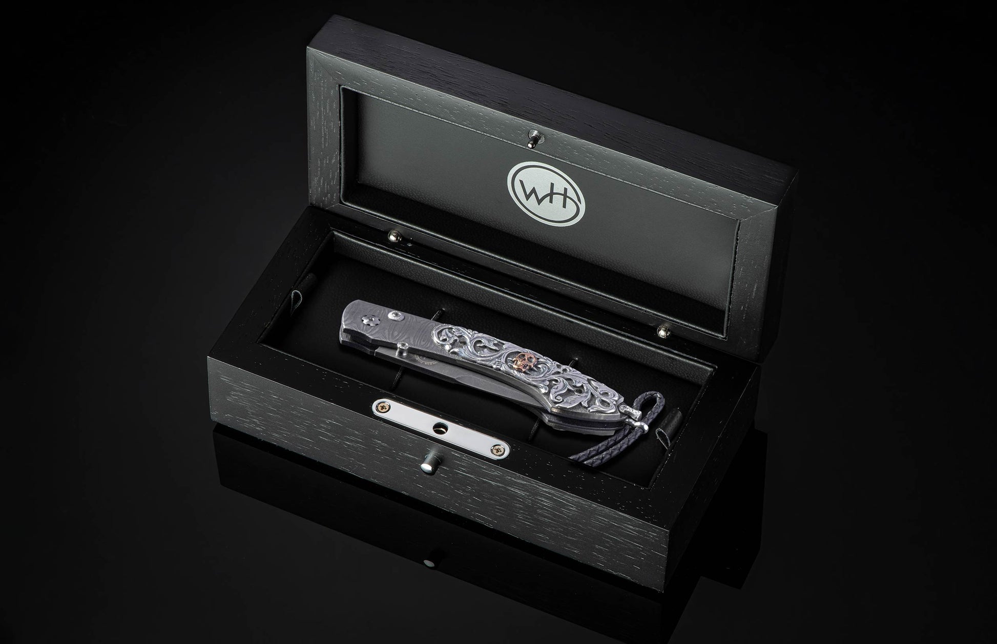 Folding knife by WH with intricate silver scrollwork and a copper skull, displayed in an open black wooden box.