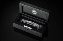 Folding knife by WH with intricate silver scrollwork and a copper skull, displayed in an open black wooden box.