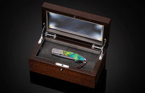 Folding knife with a vibrant green, yellow, and orange abstract handle, featuring metallic star engravings, displayed in an open wooden box.