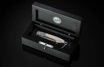 Damascus steel folding knife with contrasting layered handle and lanyard, presented in an open black wooden display box on a reflective surface.