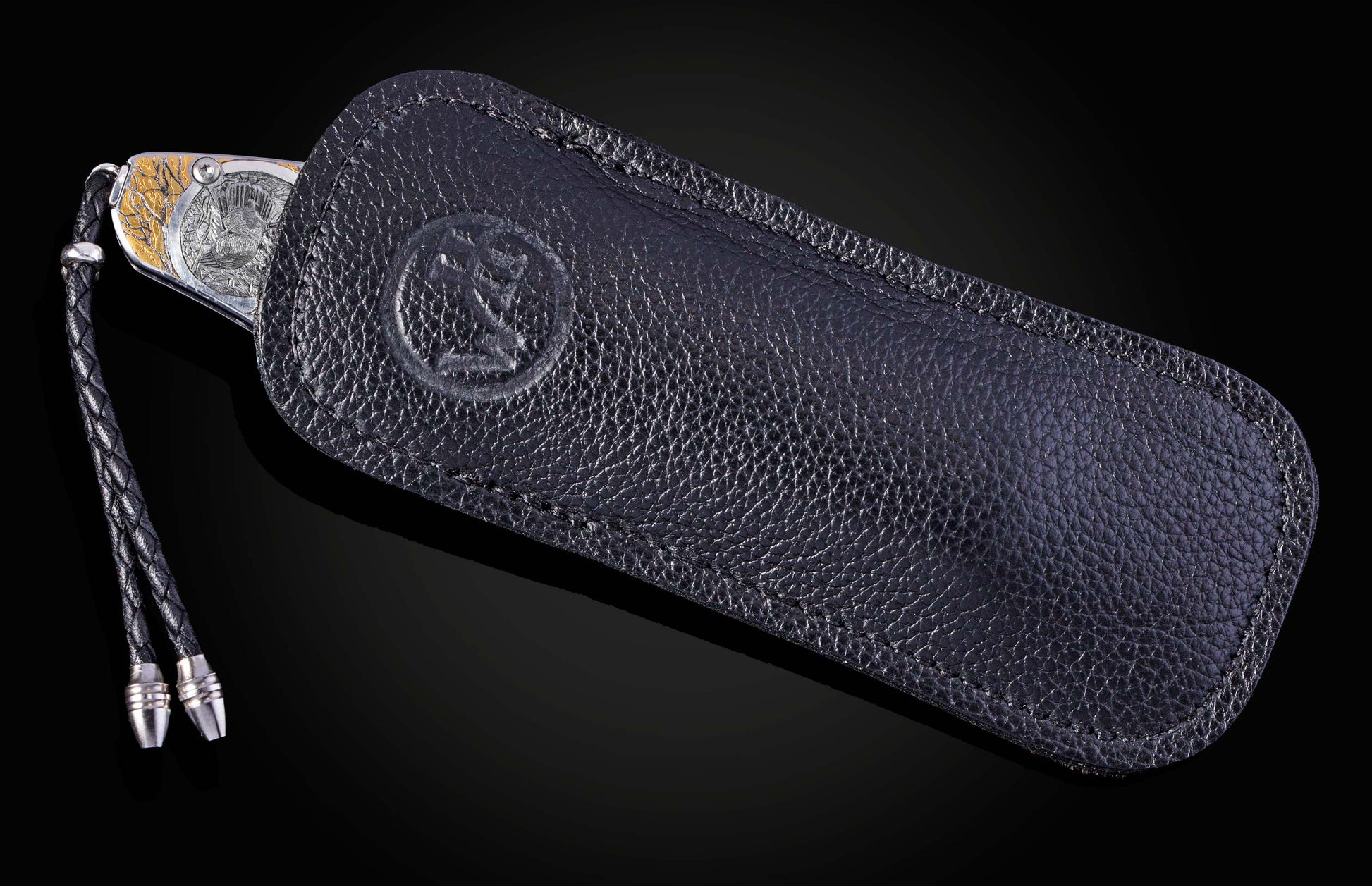 Silver and gold knife with engraved animal and tree pattern, partially in a black textured leather sheath with braided lanyard.