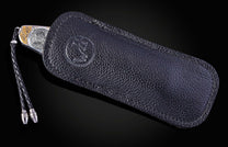 Silver and gold knife with engraved animal and tree pattern, partially in a black textured leather sheath with braided lanyard.