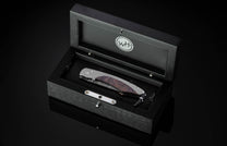 Folding knife with grey metal and textured brown handle, Damascus blade, in an open black display box.