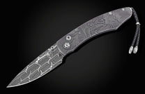 Ornate folding knife with a Damascus steel blade and engraved handle depicting a Western town scene, featuring a black leather lanyard.