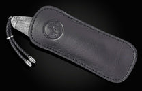 A silver-toned knife with an intricate pattern, partially sheathed in a black leather pouch with an embossed logo and a braided leather lanyard.