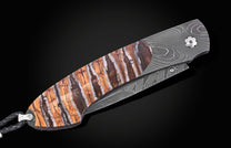 Folding knife with brown and white striped handle scales, a wavy Damascus steel blade, and a braided leather lanyard.