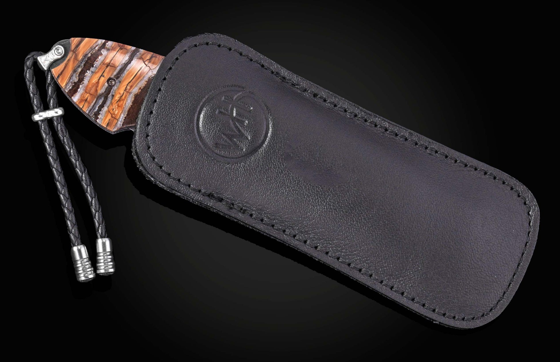 Knife partially sheathed in a black leather case, featuring a striped orange, brown, and white handle, a braided black leather lanyard with silver accents, and a circular embossed logo on the case.