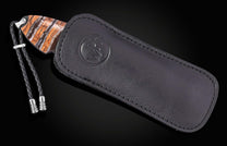 Knife partially sheathed in a black leather case, featuring a striped orange, brown, and white handle, a braided black leather lanyard with silver accents, and a circular embossed logo on the case.