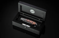 Folding knife with a patterned Damascus blade and striped natural handle, presented in an open black wooden display box.