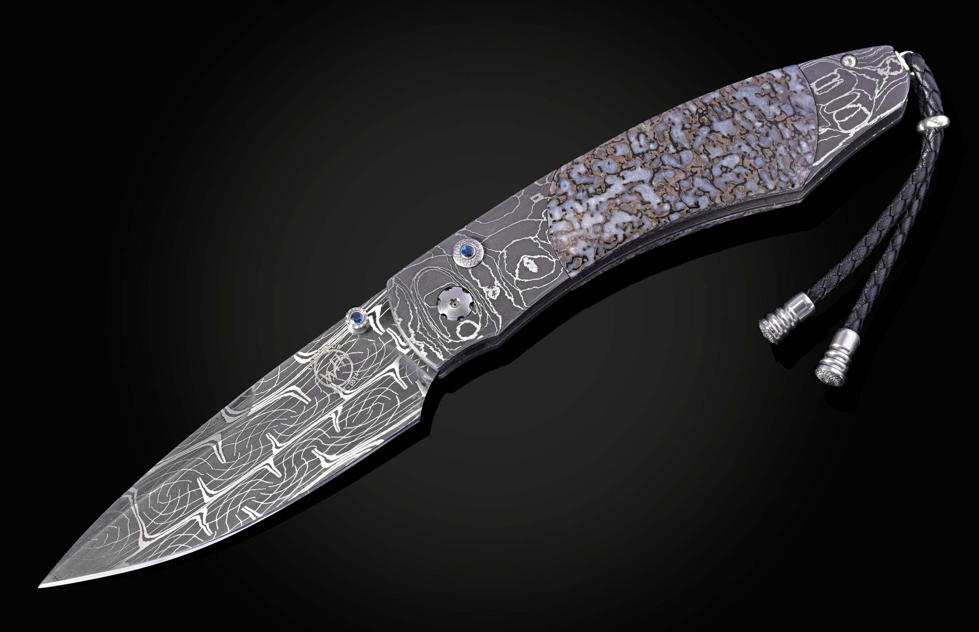 Damascus steel folding knife with an intricate handle featuring fossilized dinosaur bone, blue gemstones, and a braided leather lanyard.