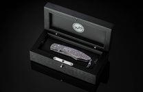 Folding knife with a damascene blade and intricate patterned handle, displayed in an open dark wood presentation box with a lanyard.