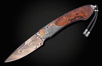 Custom folding knife with a dark burlwood handle, engraved Western-themed bolster, swirling Damascus steel blade, and a black leather lanyard.