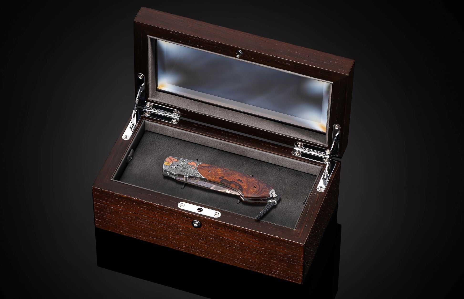 Ornate folding knife with a patterned blade and figured wooden handle, displayed in an open dark wood and glass presentation box.