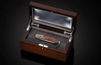 Ornate folding knife with a patterned blade and figured wooden handle, displayed in an open dark wood and glass presentation box.