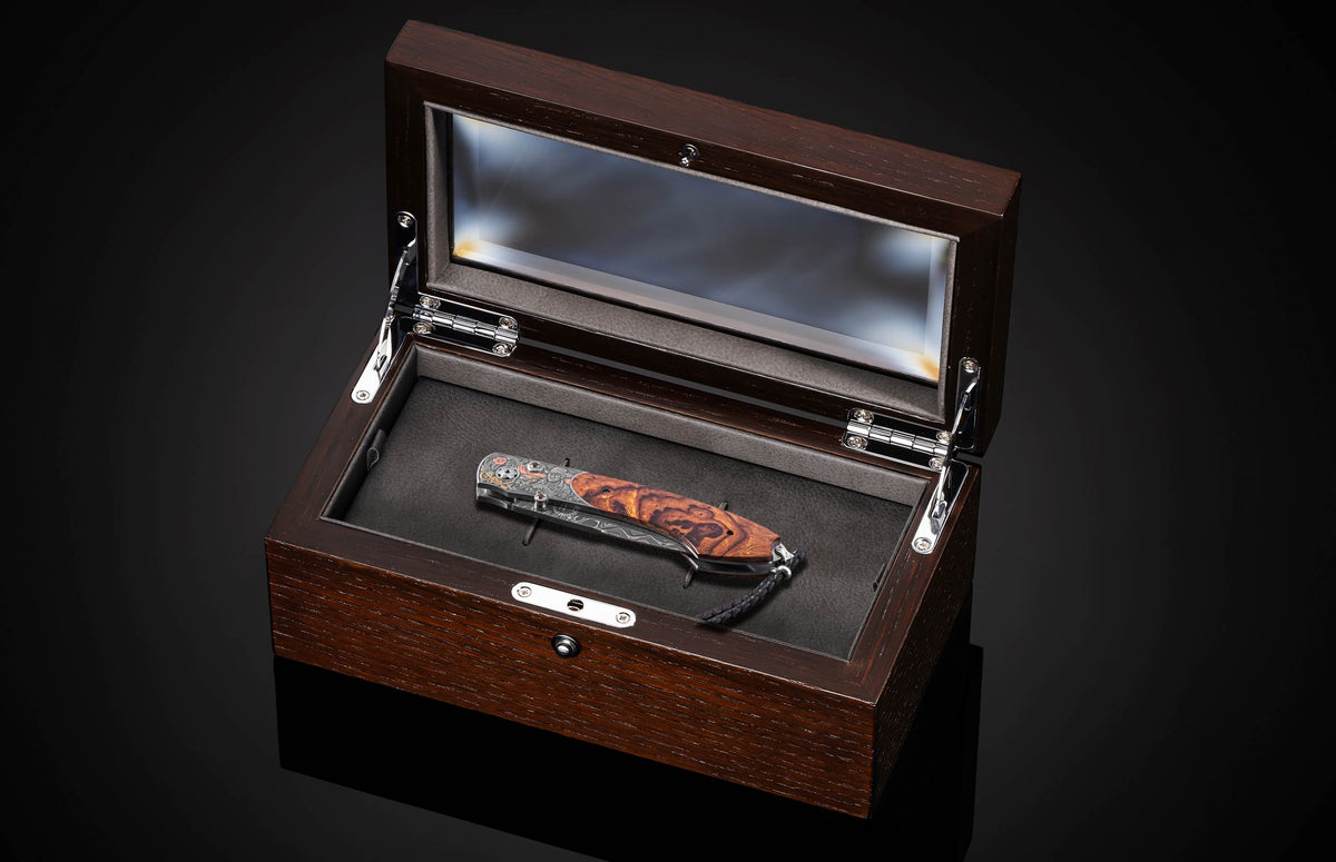 Luxury folding knife with burl wood handle and damascus blade in dark wood display case with glass lid, top-down view