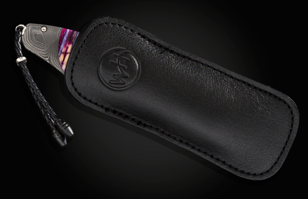 Folding knife with purple handle and damascus blade in embossed black leather sheath, shown on dark background.