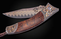 Ornate folding knife with wavy copper Damascus blade, dark burled wood handle, and silver sunburst bolsters with gems and a leather lanyard.
