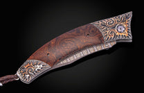 A custom folding knife with a burl wood handle, ornate silver and copper sun engravings, a Damascus pattern, and a braided leather lanyard.