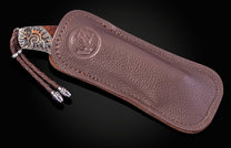 Knife with a copper sunrise handle design, partially sheathed in brown pebbled leather with an embossed logo and a braided leather thong.