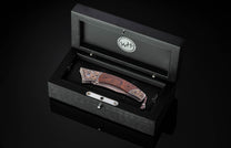 Ornate folding knife with dark wood handle, copper-toned metal, purple gem, and Damascus blade, presented in an open black wooden box.