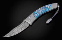 Folding knife with a patterned Damascus steel blade, a grey handle with blue urban camo inserts, blue gem accents, and a braided leather lanyard.