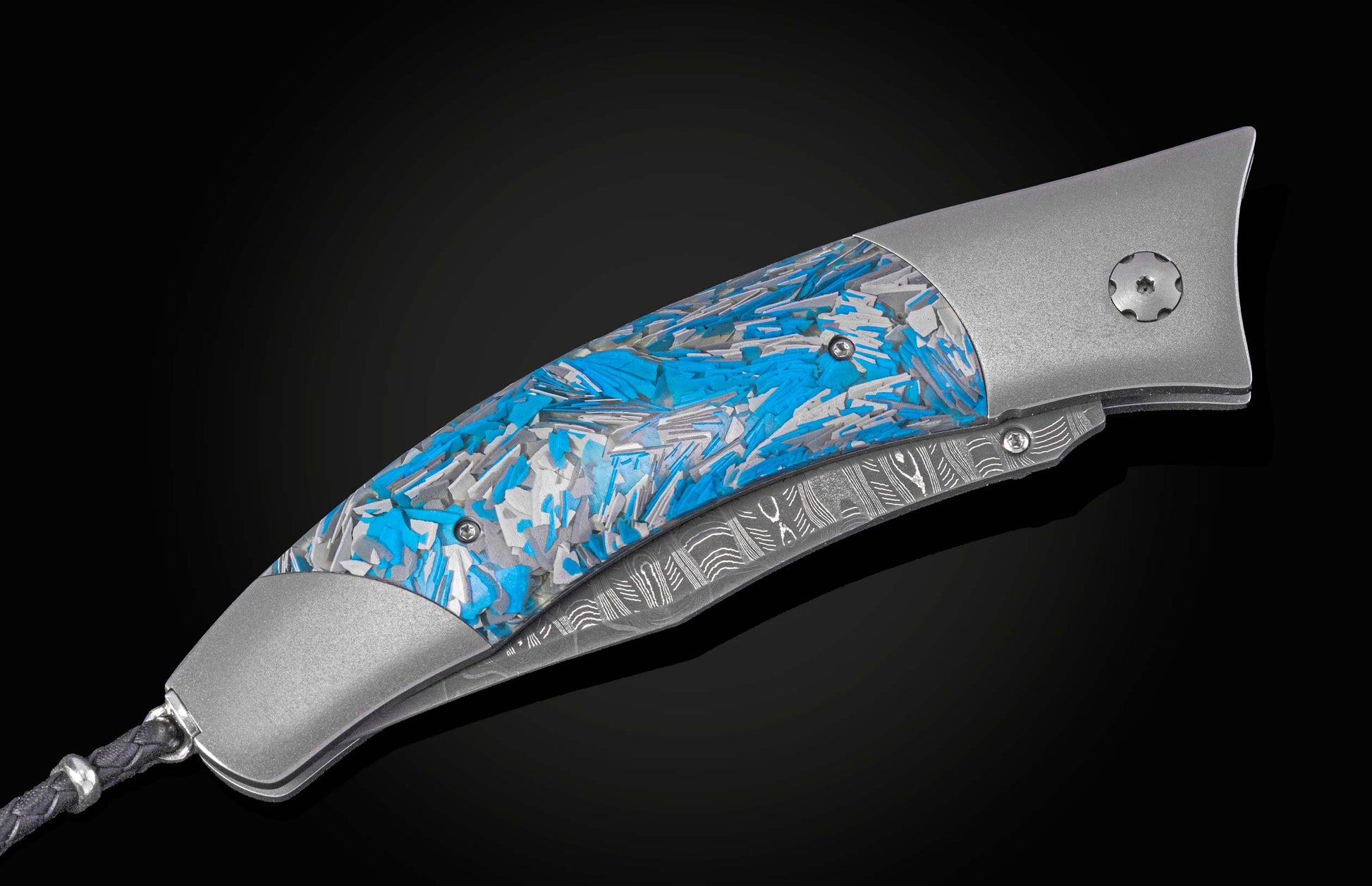 A folding knife with a textured silver handle and fragmented blue, gray, and white urban camo inlay, revealing a Damascus steel blade and braided black lanyard.