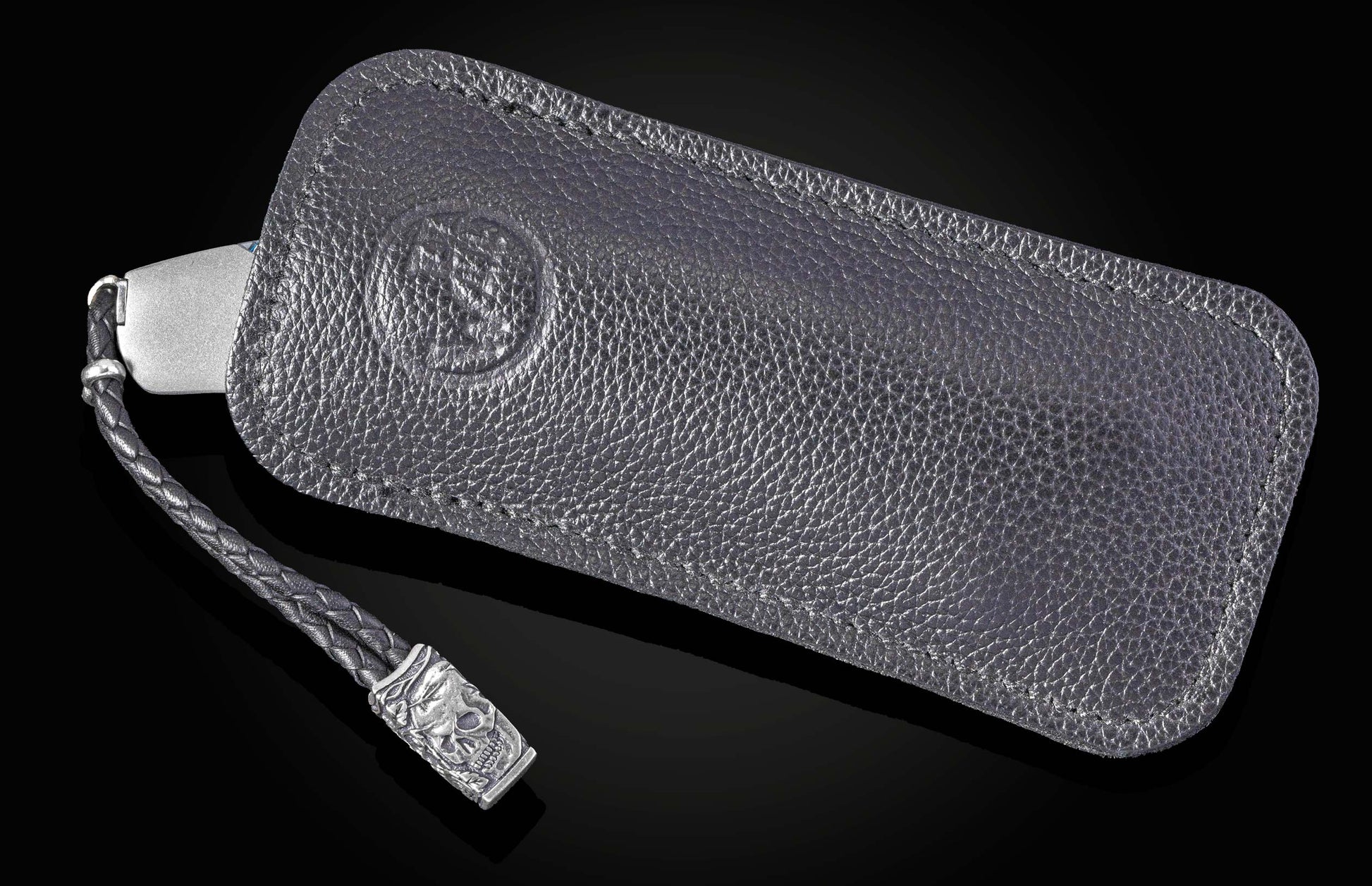 Silver-toned knife handle in a dark, textured leather case with an embossed design and a braided strap featuring a metallic skull charm.