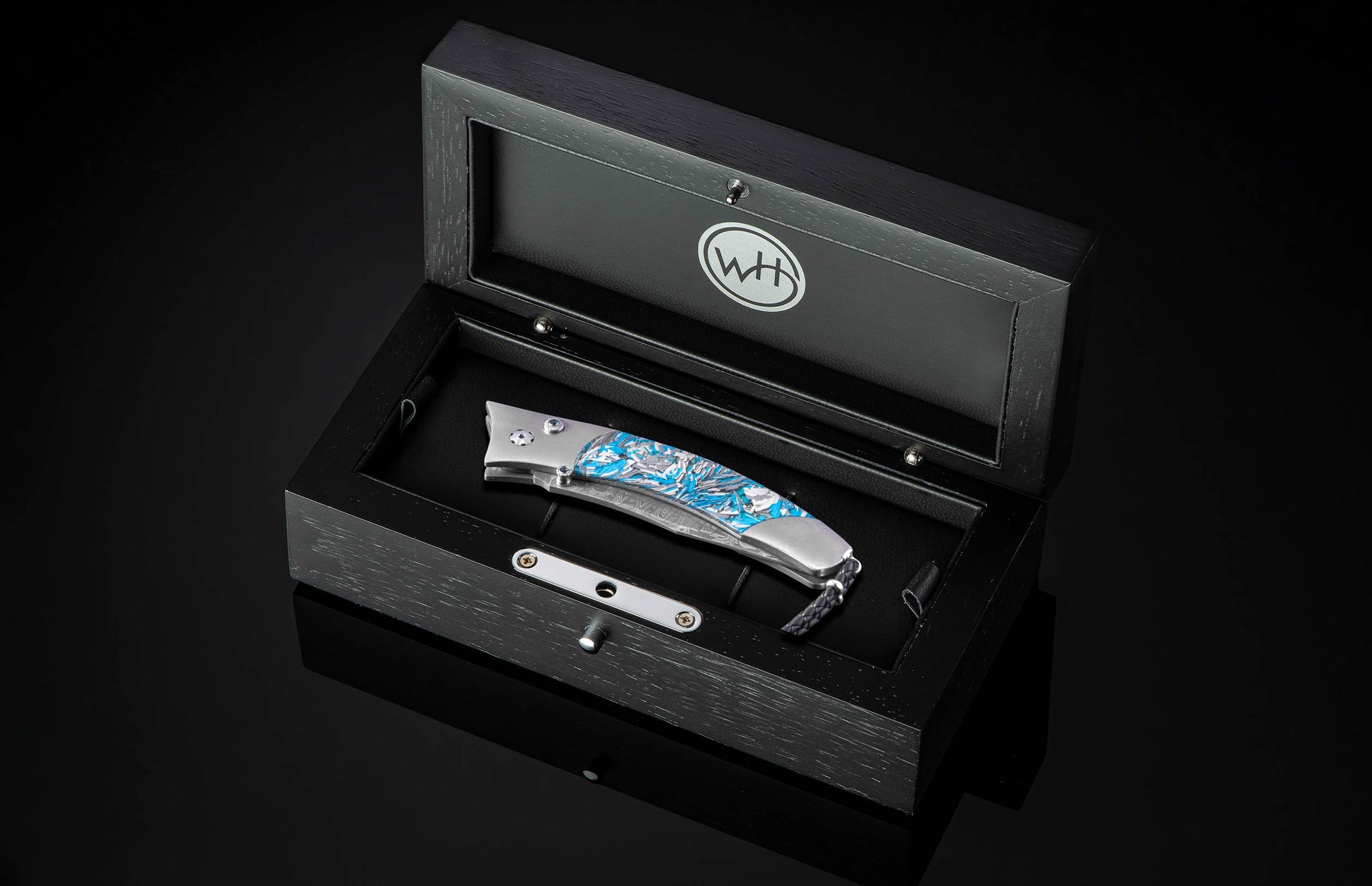 Folded pocket knife with a silver and blue Urban Camo handle, resting inside a black wooden display box with a WH logo.