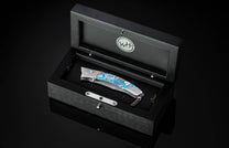 Folded pocket knife with a silver and blue Urban Camo handle, resting inside a black wooden display box with a WH logo.