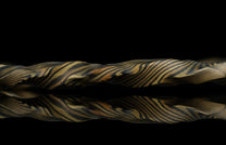 Twisted gold and black mokume-gane ring with layered wood-grain texture, reflective surface, side view on dark background