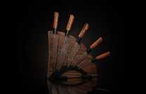 K22-BLK-W|Set of chef knives with black blades and copper rose pattern, brown wooden handles, fanned on a dark wooden block with a reflection.