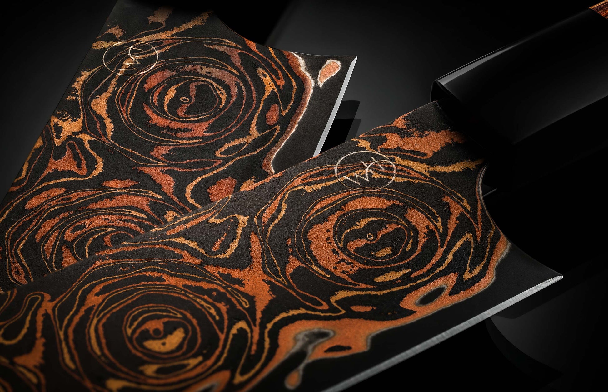 Black flare chef knives with a vibrant copper-orange swirling pattern and etched logos on the blades.