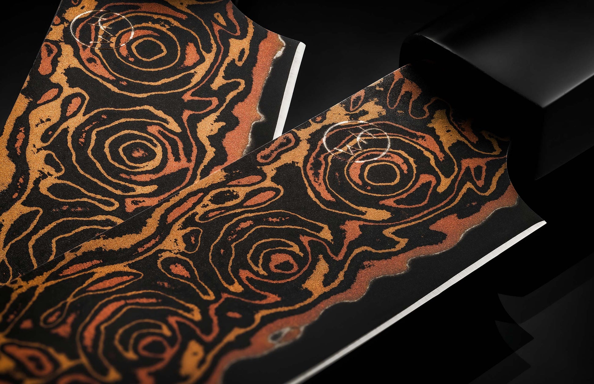 swirled pattern in knife bladeTwo black steak knives with textured orange and gold swirling patterns, a white logo, and polished blades.