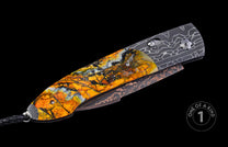 Angled auto knife with a vivid orange, yellow, and black marbled handle, a dark patterned blade with copper accents, and a black cord.